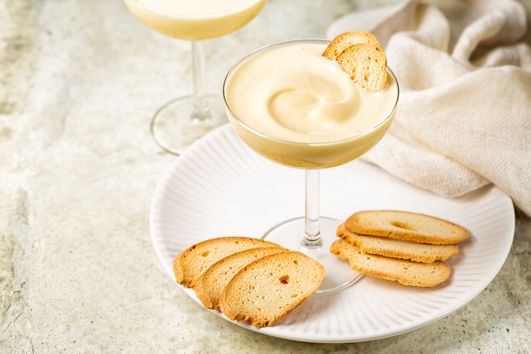 Zabaione or sabayon, or advocaat in a champagne coupe, alcoholic creamy drink  or dessert. Cream made with egg yolks, sugar, and a sweet wine Marsala or liqueur. Baicoli biscuits. White backhground, copy space.